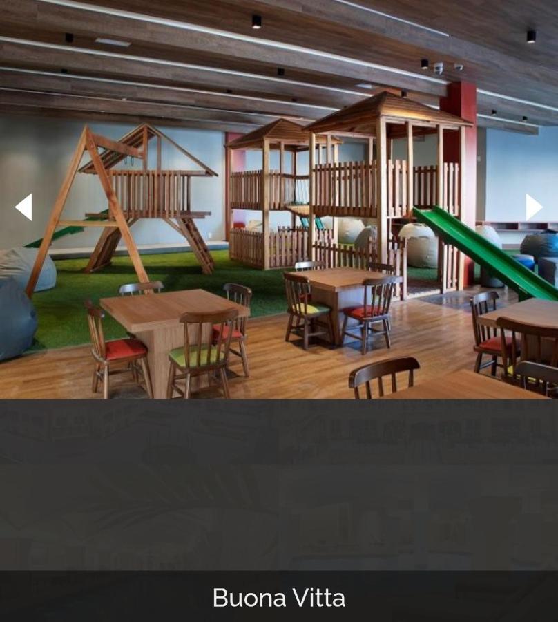 Buona Vitta Gramado Resort & Spa By Gramado Parks 외부 사진 The photo shows a play area designed for children, featuring a wooden play structure with slides and climbing areas. In the foreground, there are several small wooden tables and chairs arranged for seating. The floor is covered with green turf, resem