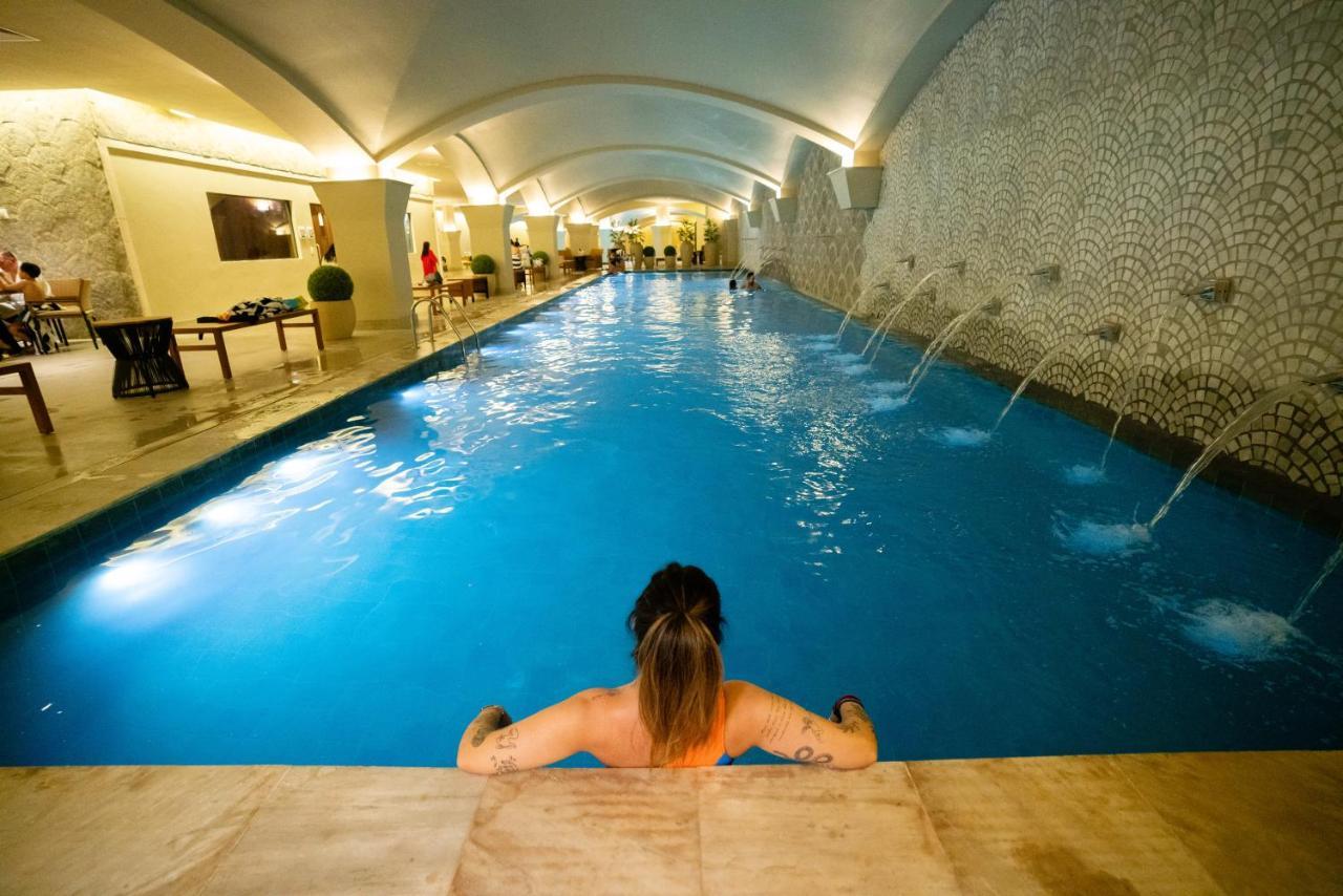 Buona Vitta Gramado Resort & Spa By Gramado Parks 외부 사진 The photo shows an indoor swimming pool with a long, narrow design. The water is a clear blue, and there are features such as fountains along one side. In the foreground, a person with long hair is sitting on the edge of the pool, looking out over th