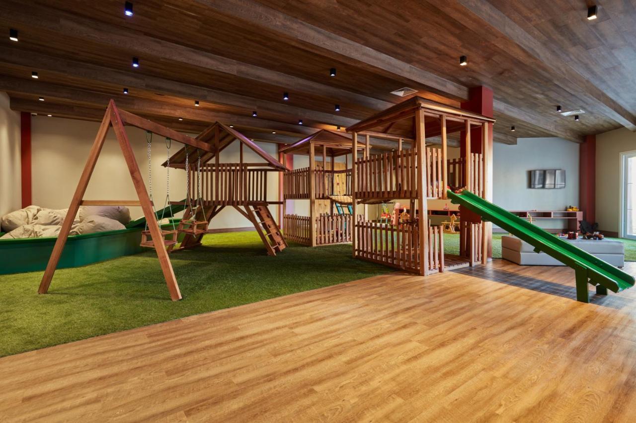 Buona Vitta Gramado Resort & Spa By Gramado Parks 외부 사진 The photo shows an indoor play area designed for children. It features a wooden play structure that includes multiple levels, slides, and a swing set. The floor is covered with wooden planks, and there is a green carpet-like area that adds a soft tou