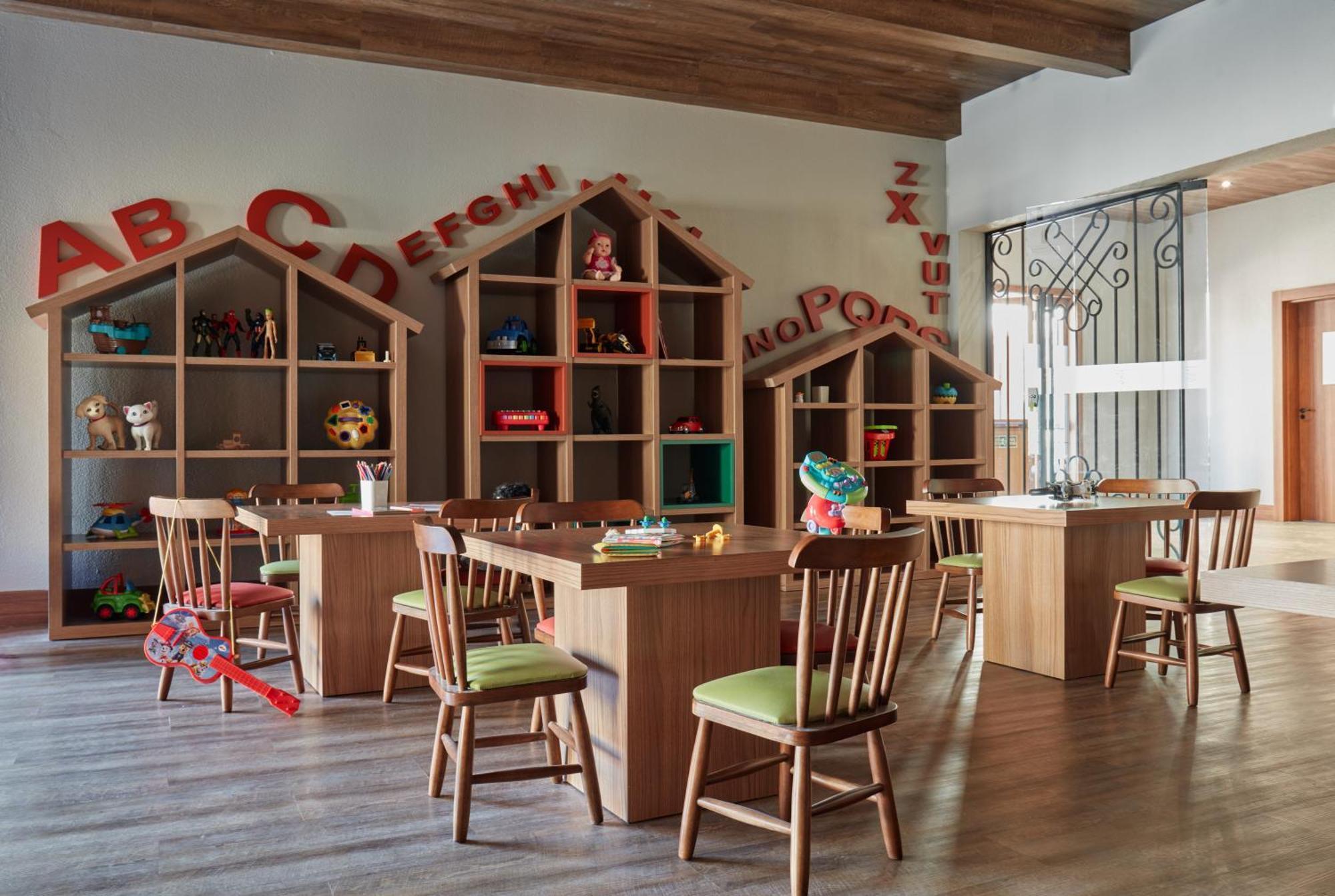 Buona Vitta Gramado Resort & Spa By Gramado Parks 외부 사진 The photo shows a classroom or playroom designed for children. It features wooden furniture, including several tables and chairs with green cushions. In the background, there are large, open shelving units shaped like houses, displaying various toys 