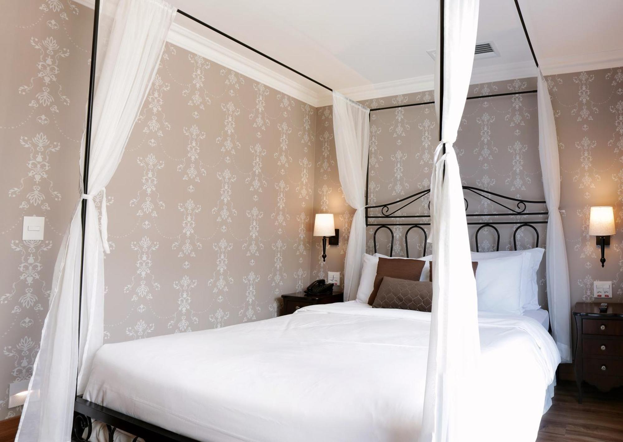 Buona Vitta Gramado Resort & Spa By Gramado Parks 외부 사진 The photo shows a elegantly decorated bedroom featuring a four-poster bed with white drapes. The bed is made with white linens and has some brown accent pillows. The walls are adorned with a subtle floral pattern, and there are two bedside lamps prov