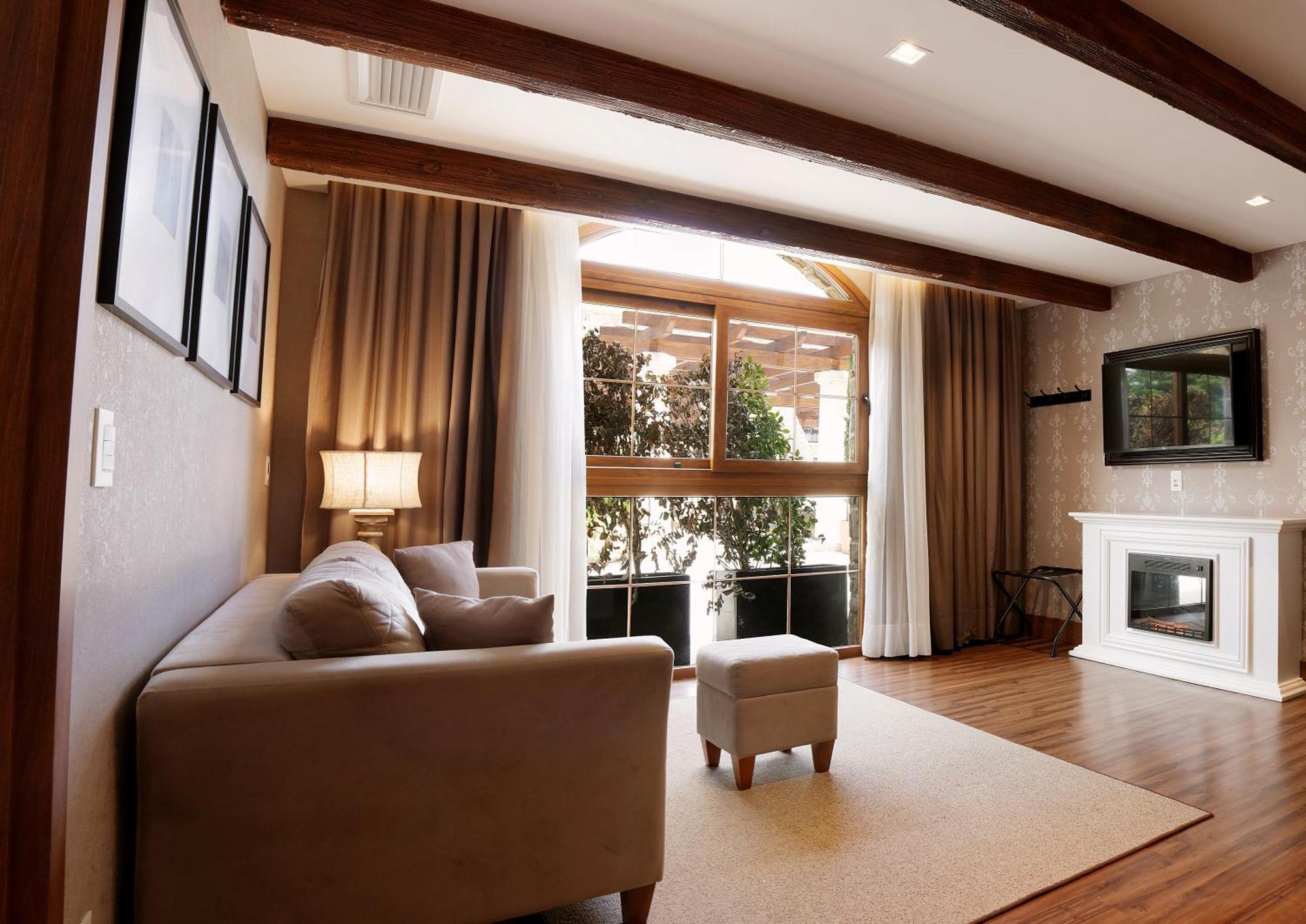 Buona Vitta Gramado Resort & Spa By Gramado Parks 외부 사진 The photo depicts a cozy living room setting. It features a light-colored couch, a small ottoman, and a floor lamp beside the couch. Large windows are covered with curtains, allowing natural light to illuminate the space. In the background, there is 