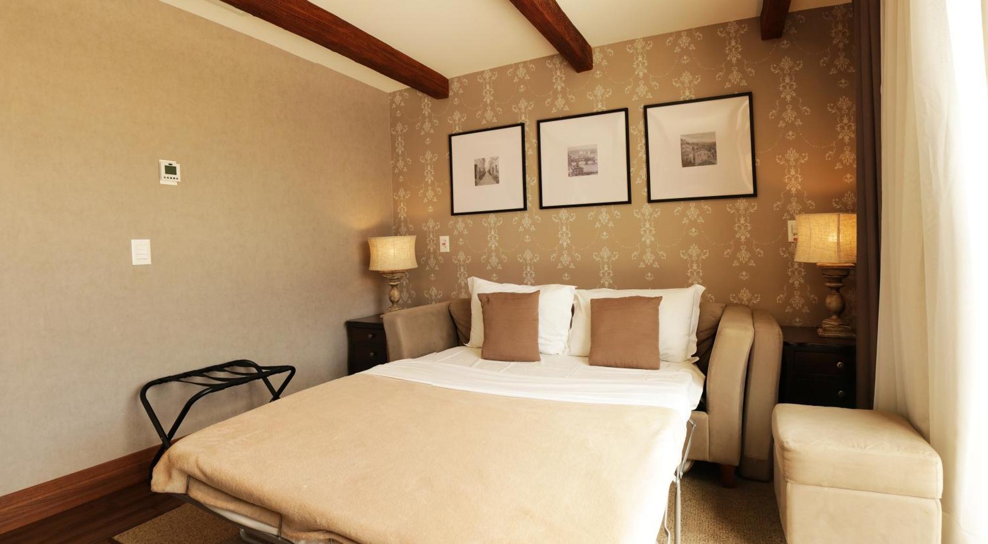 Buona Vitta Gramado Resort & Spa By Gramado Parks 외부 사진 The photo shows a cozy bedroom with a neutral color palette. There is a bed with white linens and two brown decorative pillows. On either side of the bed, there are bedside tables with lamps. The walls feature a subtle floral pattern, and three frame