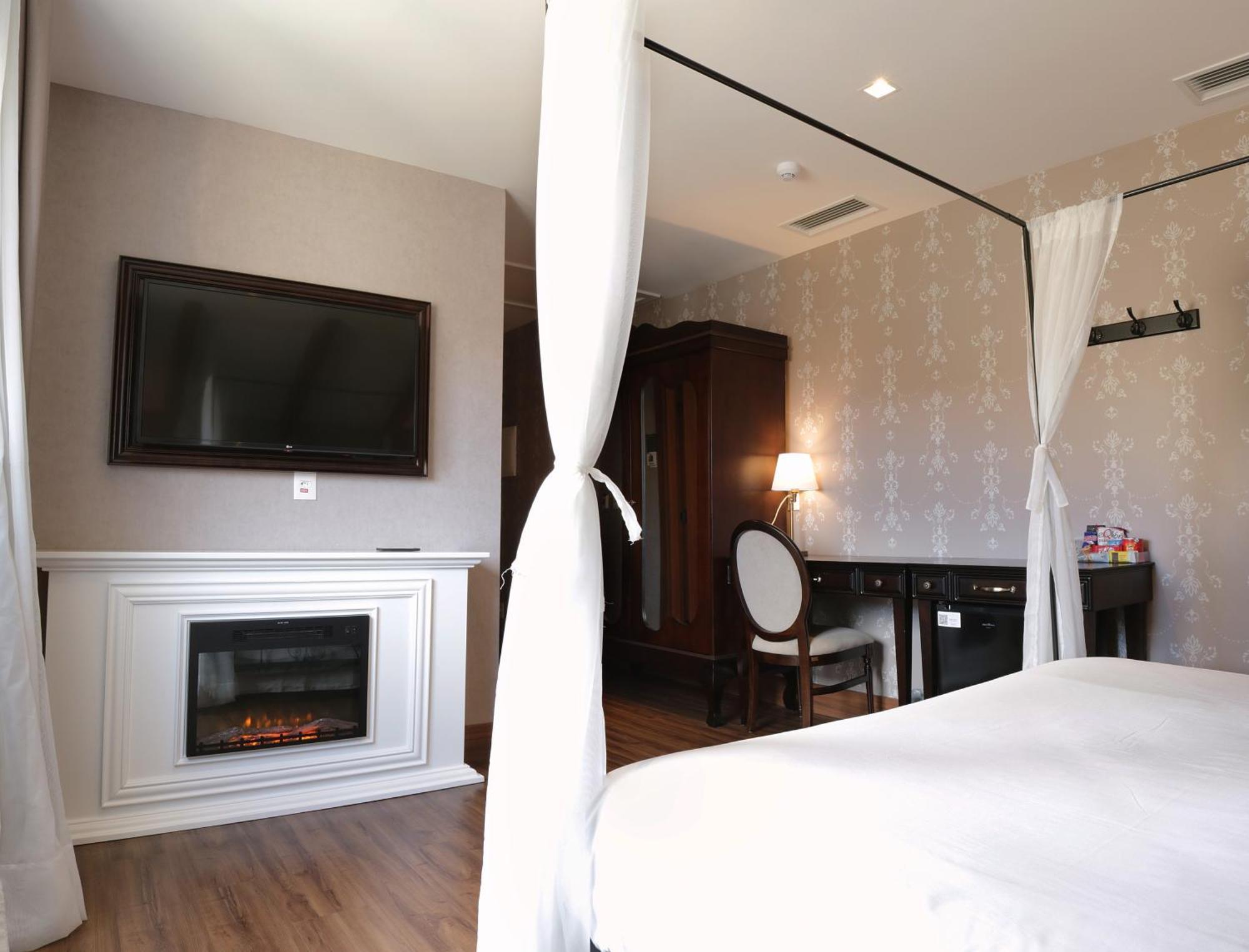 Buona Vitta Gramado Resort & Spa By Gramado Parks 외부 사진 The photo shows a cozy hotel room with elegant decor. There is a white bed with a canopy, and a modern television mounted on the wall across from it. To the side, a fireplace is built into a white mantel, adding a warm touch to the space. There's a w