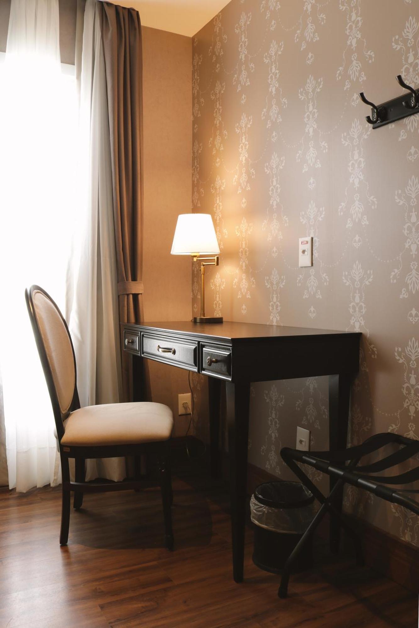 Buona Vitta Gramado Resort & Spa By Gramado Parks 외부 사진 The photo shows a small desk set against a wall with a decorative pattern. The desk is black with a few drawers and has a small lamp on it. Next to the desk, there is a chair with a light-colored cushion. Soft light is coming through a window, castin