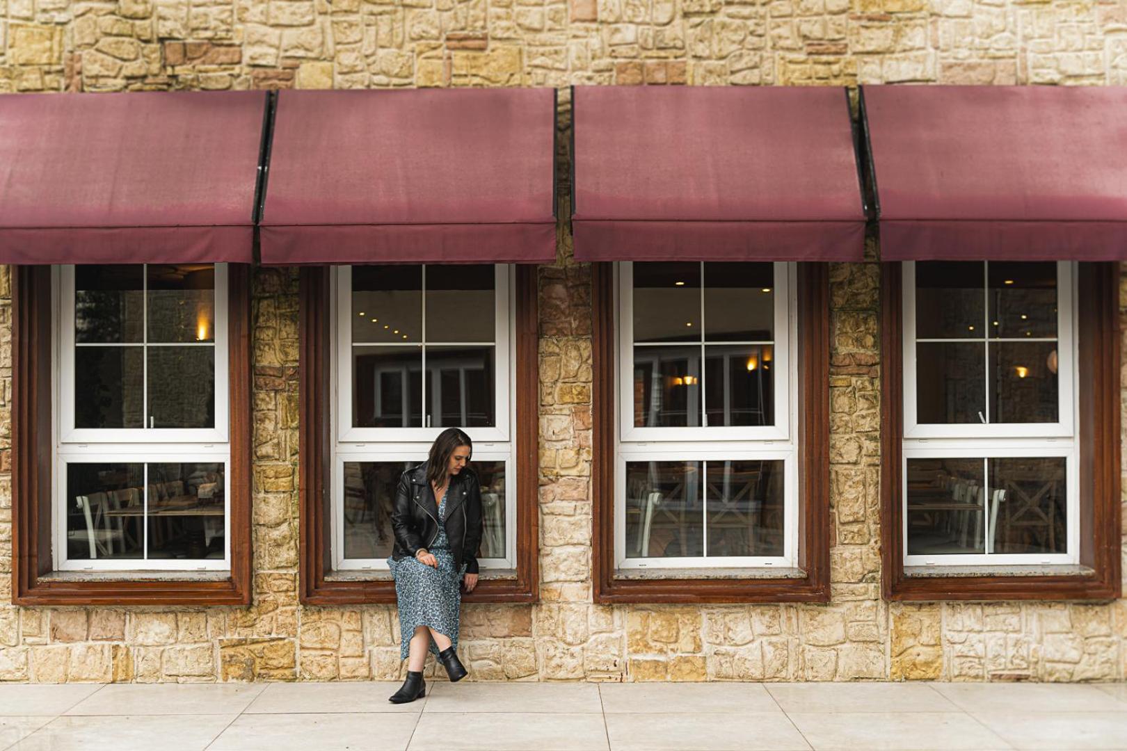 Buona Vitta Gramado Resort & Spa By Gramado Parks 외부 사진 The photo shows a woman standing against a stone wall with three large windows in front of her, each framed with wooden borders. The windows have maroon awnings above them. The woman is dressed in a long skirt and leather jacket, leaning slightly aga